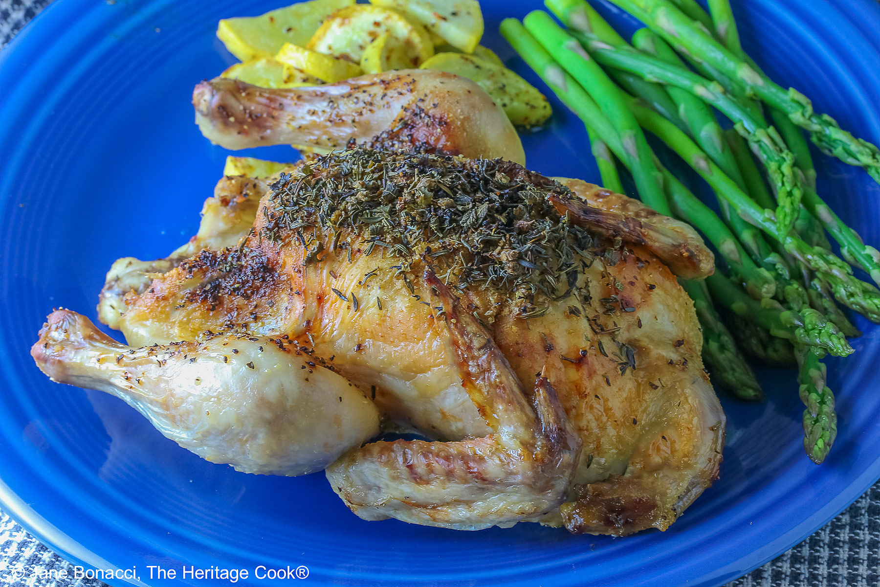A Roasted Game Hen with Herb Butter for Valentine's Day on a deep blue plate with sauteed yellow squash and steamed asparagus, sitting on a checked cloth © 2025 Jane Bonacci, The Heritage Cook. 