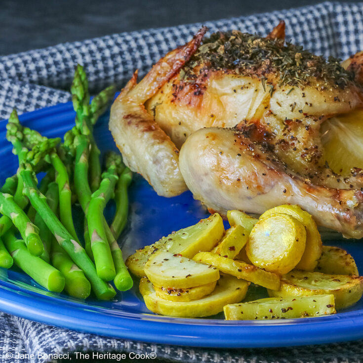 A Roasted Game Hen with Herb Butter for Valentine's Day on a deep blue plate with sauteed yellow squash and steamed asparagus, sitting on a checked cloth © 2025 Jane Bonacci, The Heritage Cook.