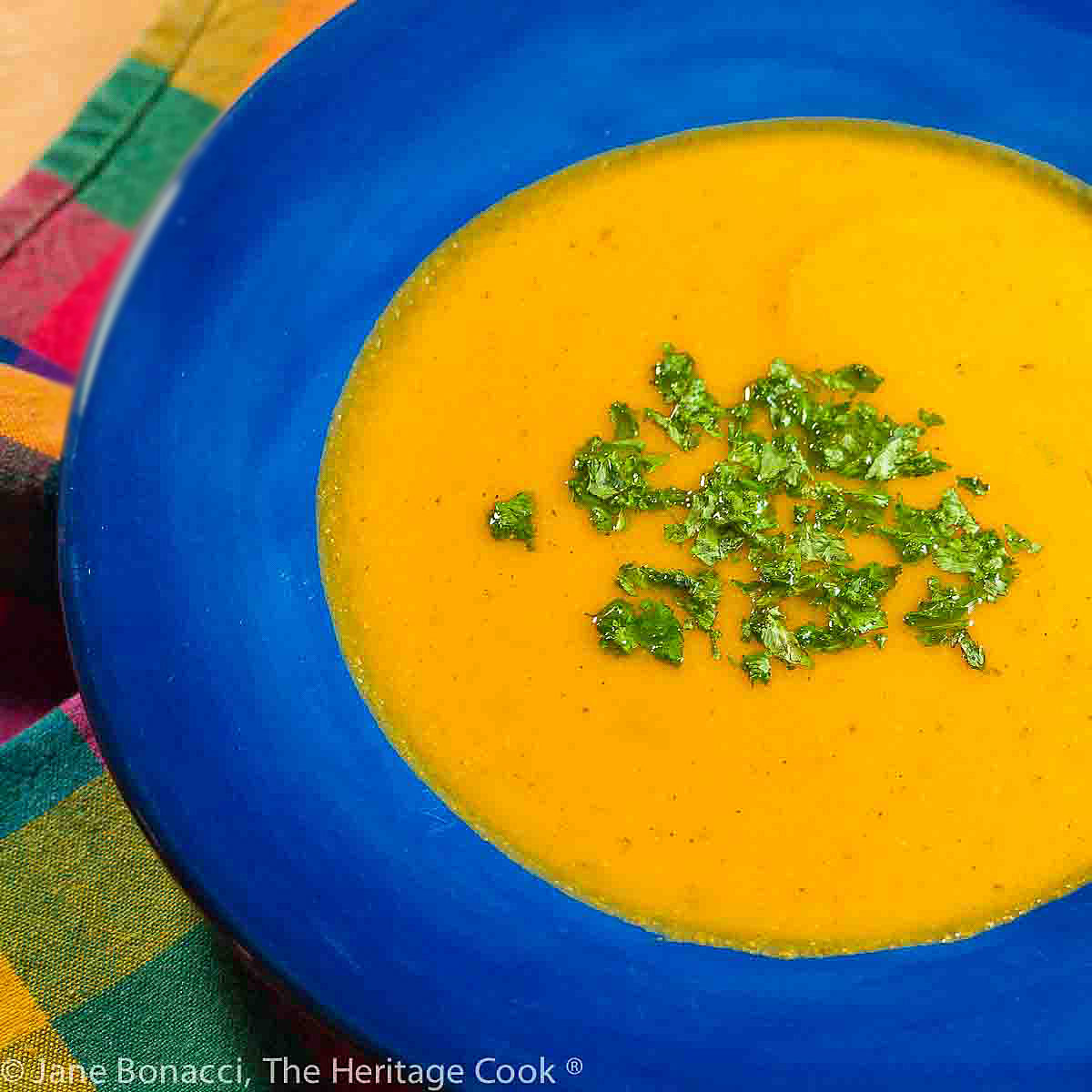 Deep orange golden soup in a dark blue bowl on a multicolored cloth with green chopped herbs on top. Enjoy this Roasted Carrot Sweet Potato Soup (Gluten-Free; Dairy-Free); 2024 Jane Bonacci, The Heritage Cook. 