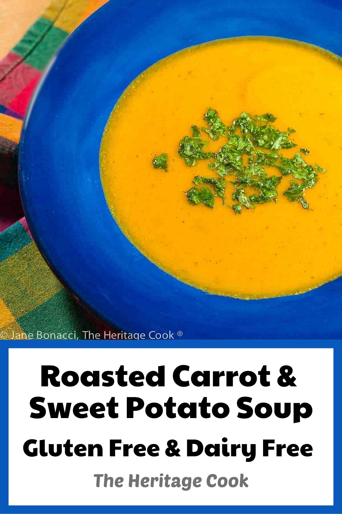 Deep orange golden soup in a dark blue bowl on a multicolored cloth with green chopped herbs on top. Enjoy this Roasted Carrot Sweet Potato Soup (Gluten-Free; Dairy-Free); 2024 Jane Bonacci, The Heritage Cook. 
