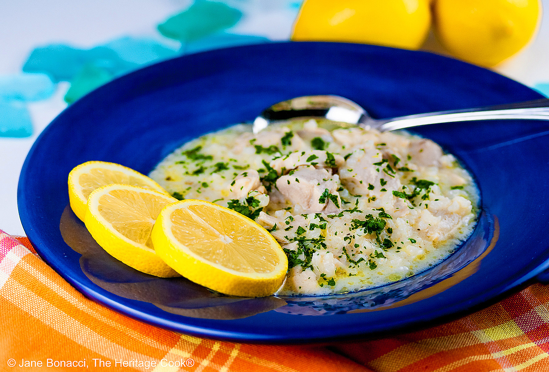 Avgolemono – Greek Lemon Chicken Soup – in a very full deep blue bowl with 3 bright yellow lemon slices on the rim, with a spoon in the bowl or next to it, sitting on an orange and yellow cloth © 2026 Jane Bonacci, The Heritage Cook.