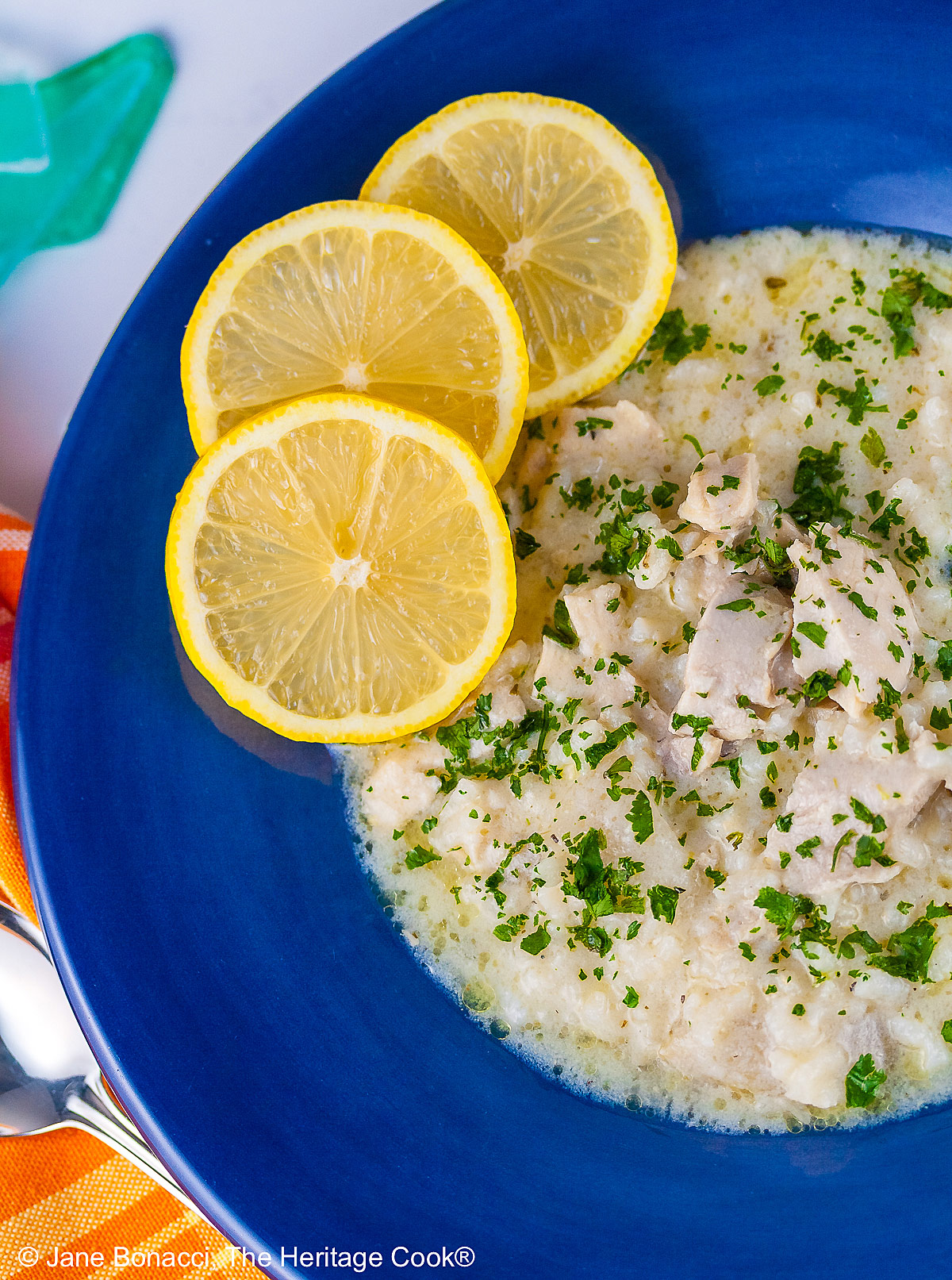 Avgolemono – Greek Lemon Chicken Soup – in a very full deep blue bowl with 3 bright yellow lemon slices on the rim, with a spoon in the bowl or next to it, sitting on an orange and yellow cloth © 2026 Jane Bonacci, The Heritage Cook.