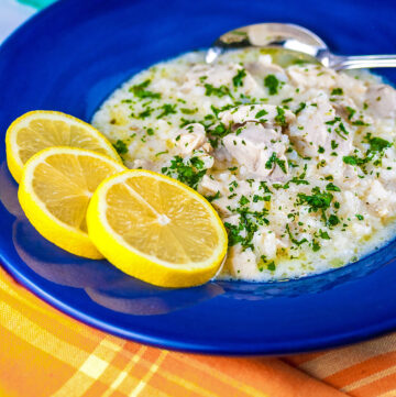 Avgolemono – Greek Lemon Chicken Soup – in a very full deep blue bowl with 3 bright yellow lemon slices on the rim, with a spoon in the bowl or next to it, sitting on an orange and yellow cloth © 2026 Jane Bonacci, The Heritage Cook.
