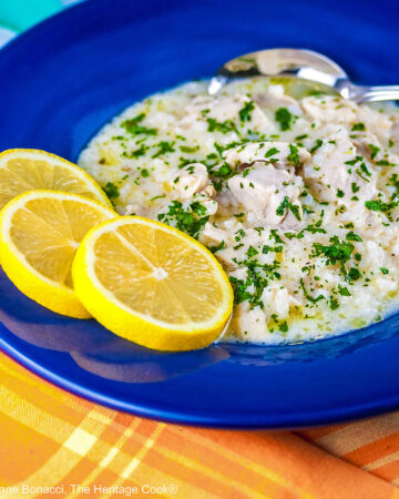 Avgolemono – Greek Lemon Chicken Soup – in a very full deep blue bowl with 3 bright yellow lemon slices on the rim, with a spoon in the bowl or next to it, sitting on an orange and yellow cloth © 2026 Jane Bonacci, The Heritage Cook.