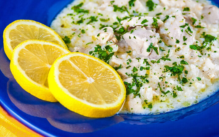 Avgolemono – Greek Lemon Chicken Soup – in a very full deep blue bowl with 3 bright yellow lemon slices on the rim, with a spoon in the bowl or next to it, sitting on an orange and yellow cloth © 2026 Jane Bonacci, The Heritage Cook.