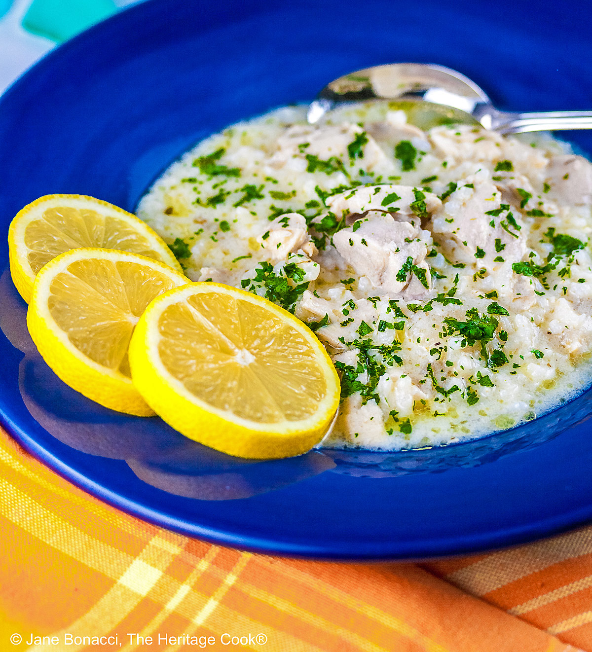 Avgolemono – Greek Lemon Chicken Soup – in a very full deep blue bowl with 3 bright yellow lemon slices on the rim, with a spoon in the bowl or next to it, sitting on an orange and yellow cloth © 2026 Jane Bonacci, The Heritage Cook.