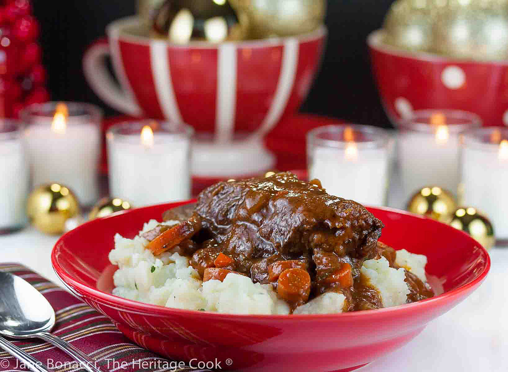 Red Wine Braised Short Ribs and carrots nestled into a red bowl filled with garlic mashed potatoes, in front of festive candles and Christmas decorations © 2023 Jane Bonacci, The Heritage Cook.