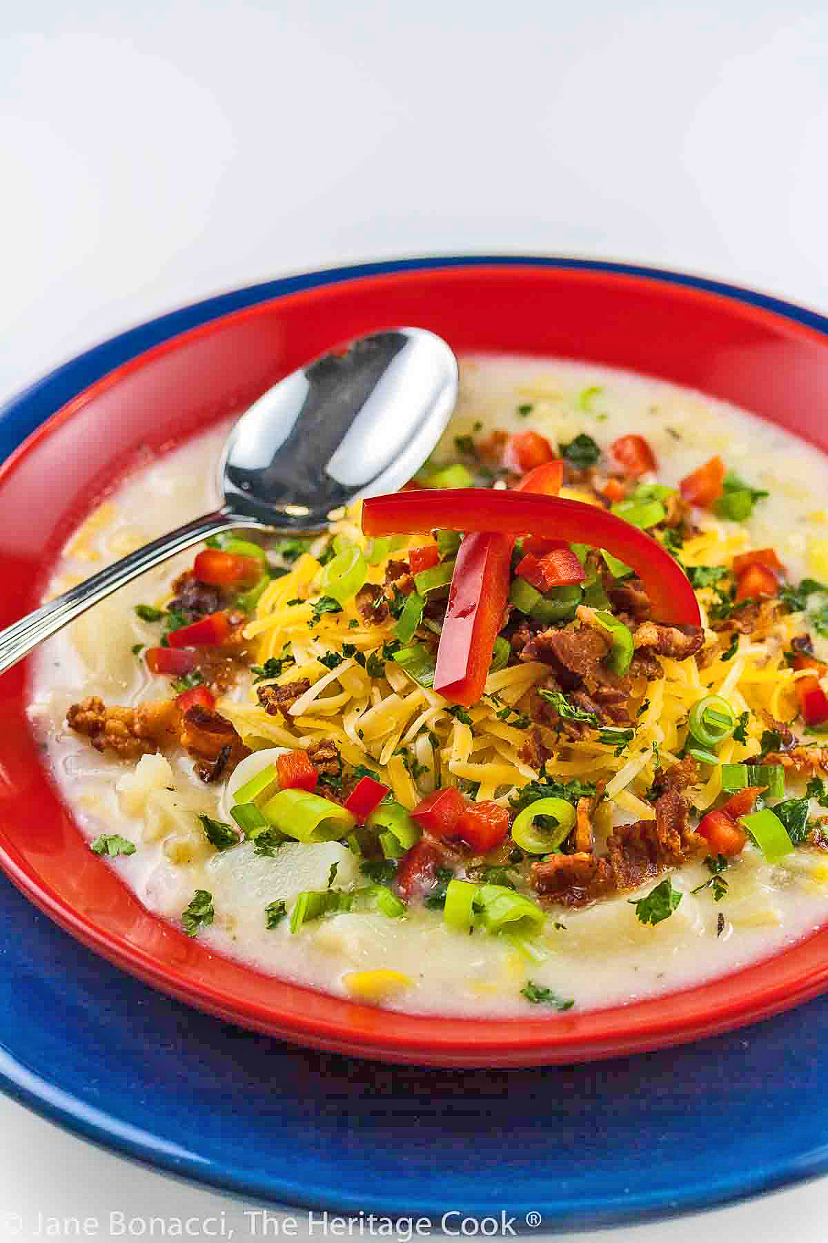 Light colored soup in a red bowl sitting on a dark blue plate, topped with shredded cheese, red bell peppers, bacon, green onions © 2024 Jane Bonacci, The Heritage Cook. 