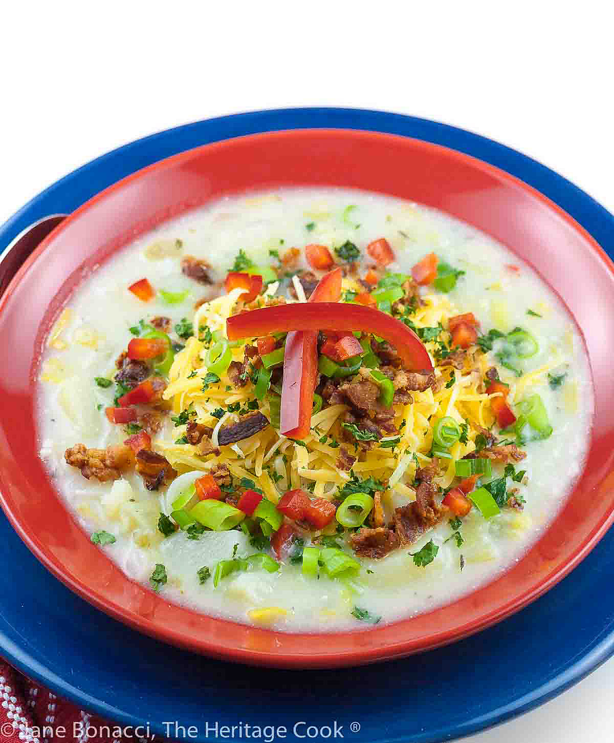 Light colored soup in a red bowl sitting on a dark blue plate, topped with shredded cheese, red bell peppers, bacon, green onions © 2024 Jane Bonacci, The Heritage Cook. 