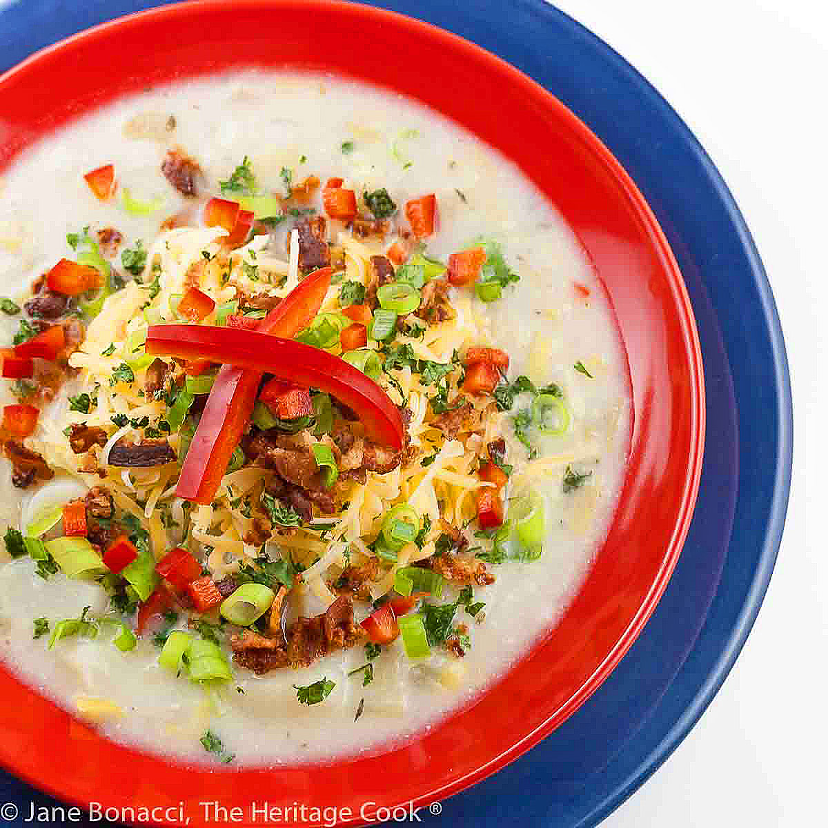 Light colored soup in a red bowl sitting on a dark blue plate, topped with shredded cheese, red bell peppers, bacon, green onions © 2024 Jane Bonacci, The Heritage Cook.