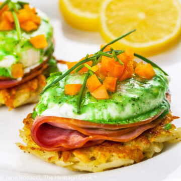 Potato pancakes topped with a slice of ham, a poached egg, basil hollandaise sauce, finely chopped orange bell peppers, and chives; Irish Hash Browns Benedict with Basil Hollandaise; © 2023 Jane Bonacci, The Heritage Cook.