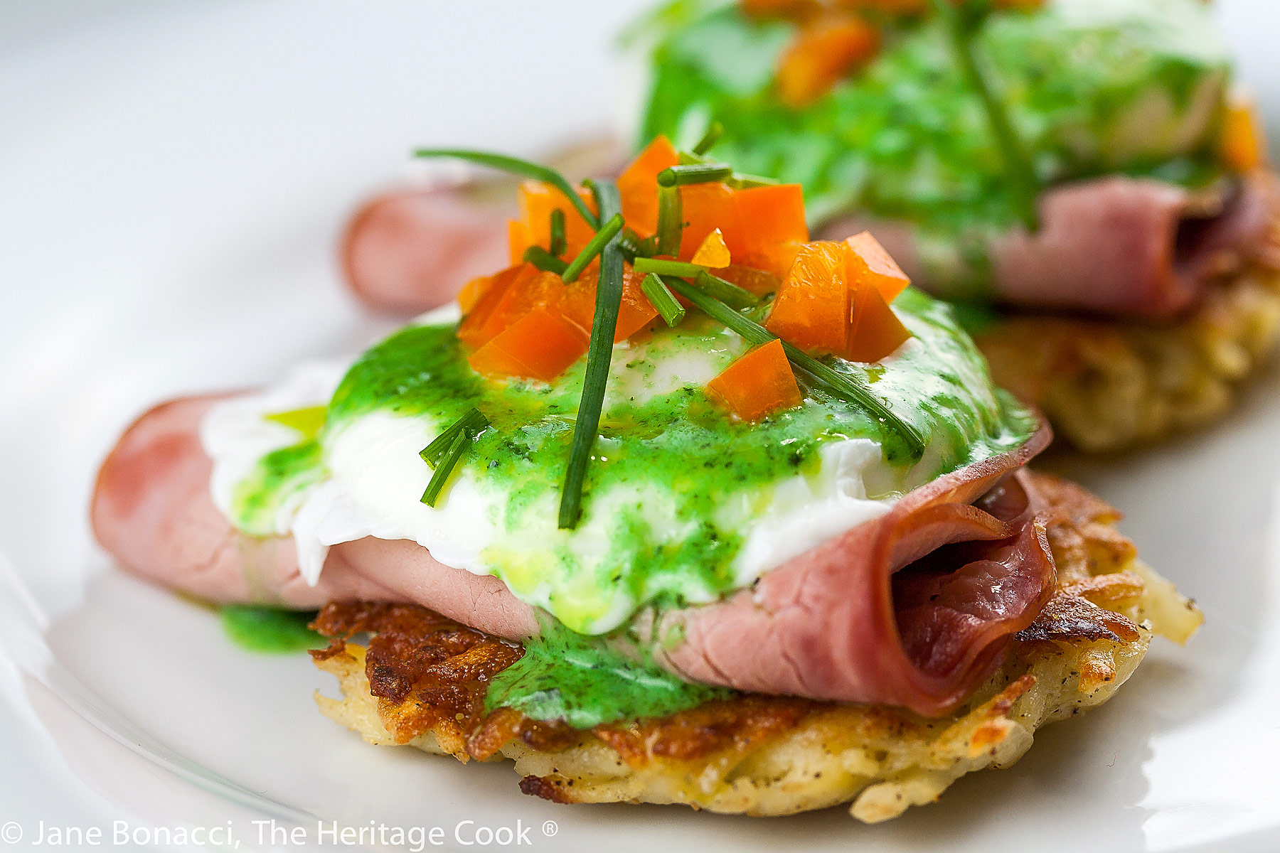 Potato pancakes topped with a slice of ham, a poached egg, basil hollandaise sauce, finely chopped orange bell peppers, and chives; Irish Hash Browns Benedict with Basil Hollandaise; © 2023 Jane Bonacci, The Heritage Cook. 