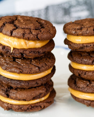 Stacks of Chocolate Peanut Butter Sandwich Cookies on a white plate with a scalloped blue edge, on a dark blue background some with a bowl of the filling to the side and a cloth with the Eiffel Tower on it behind © 2026 Jane Bonacci, The Heritage Cook.