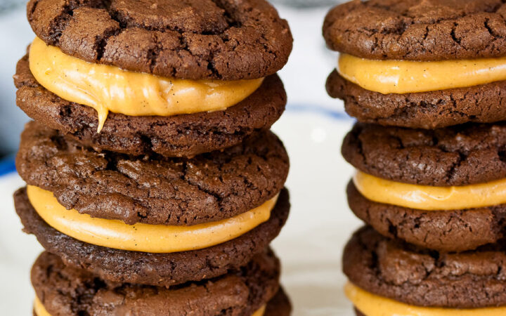 Stacks of Chocolate Peanut Butter Sandwich Cookies on a white plate with a scalloped blue edge, on a dark blue background some with a bowl of the filling to the side and a cloth with the Eiffel Tower on it behind © 2026 Jane Bonacci, The Heritage Cook.