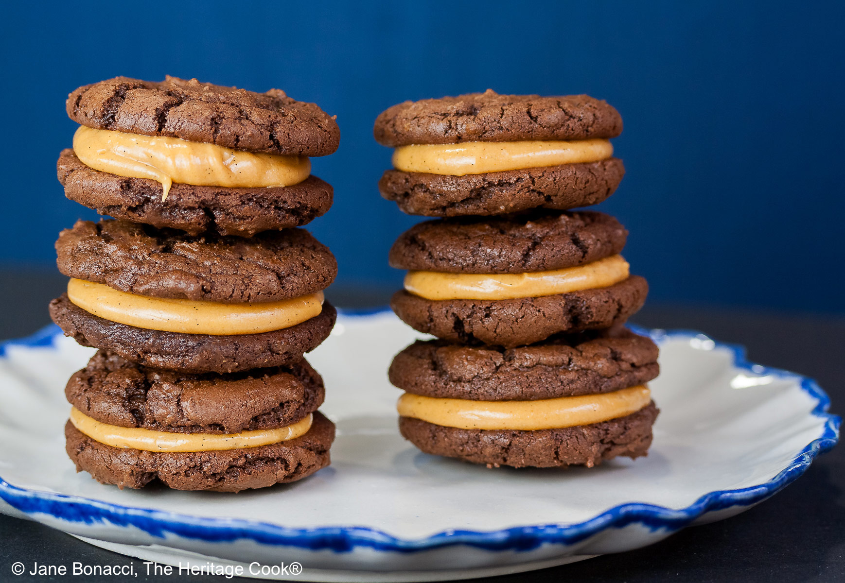 Stacks of Chocolate Peanut Butter Sandwich Cookies on a white plate with a scalloped blue edge, on a dark blue background some with a bowl of the filling to the side and a cloth with the Eiffel Tower on it behind © 2026 Jane Bonacci, The Heritage Cook.