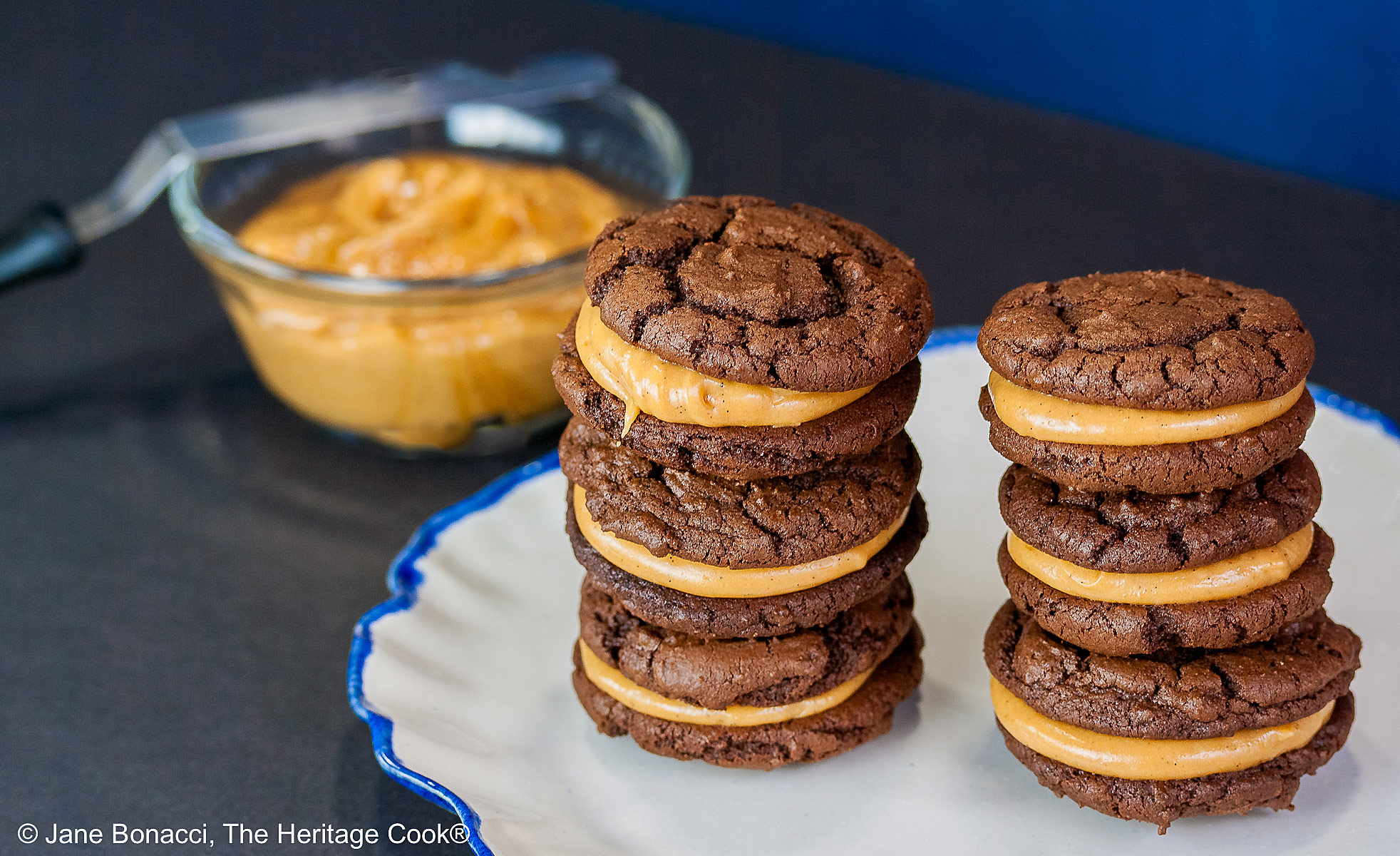 Stacks of Chocolate Peanut Butter Sandwich Cookies on a white plate with a scalloped blue edge, on a dark blue background some with a bowl of the filling to the side and a cloth with the Eiffel Tower on it behind © 2026 Jane Bonacci, The Heritage Cook.