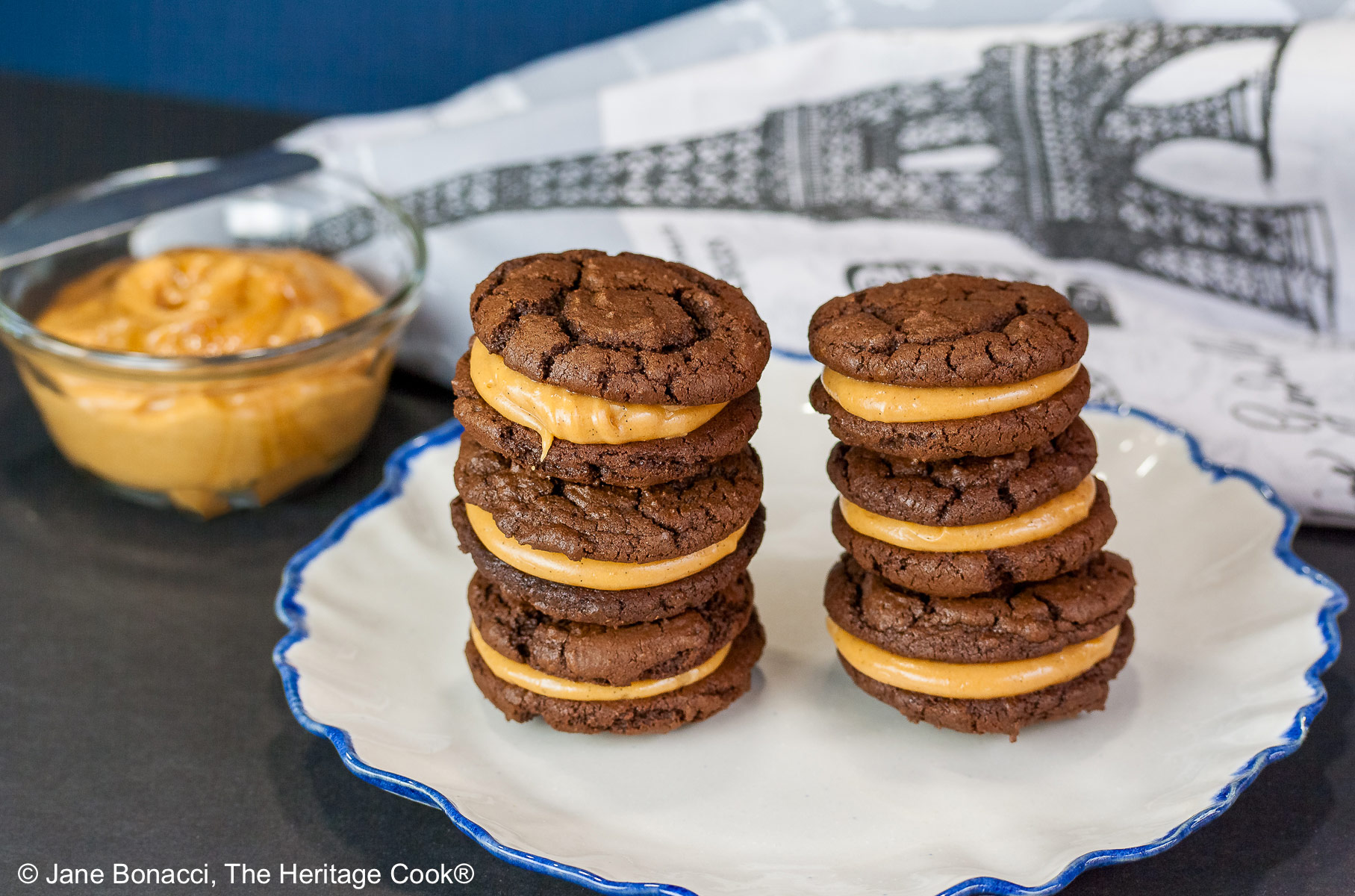 Stacks of Chocolate Peanut Butter Sandwich Cookies on a white plate with a scalloped blue edge, on a dark blue background some with a bowl of the filling to the side and a cloth with the Eiffel Tower on it behind © 2026 Jane Bonacci, The Heritage Cook.