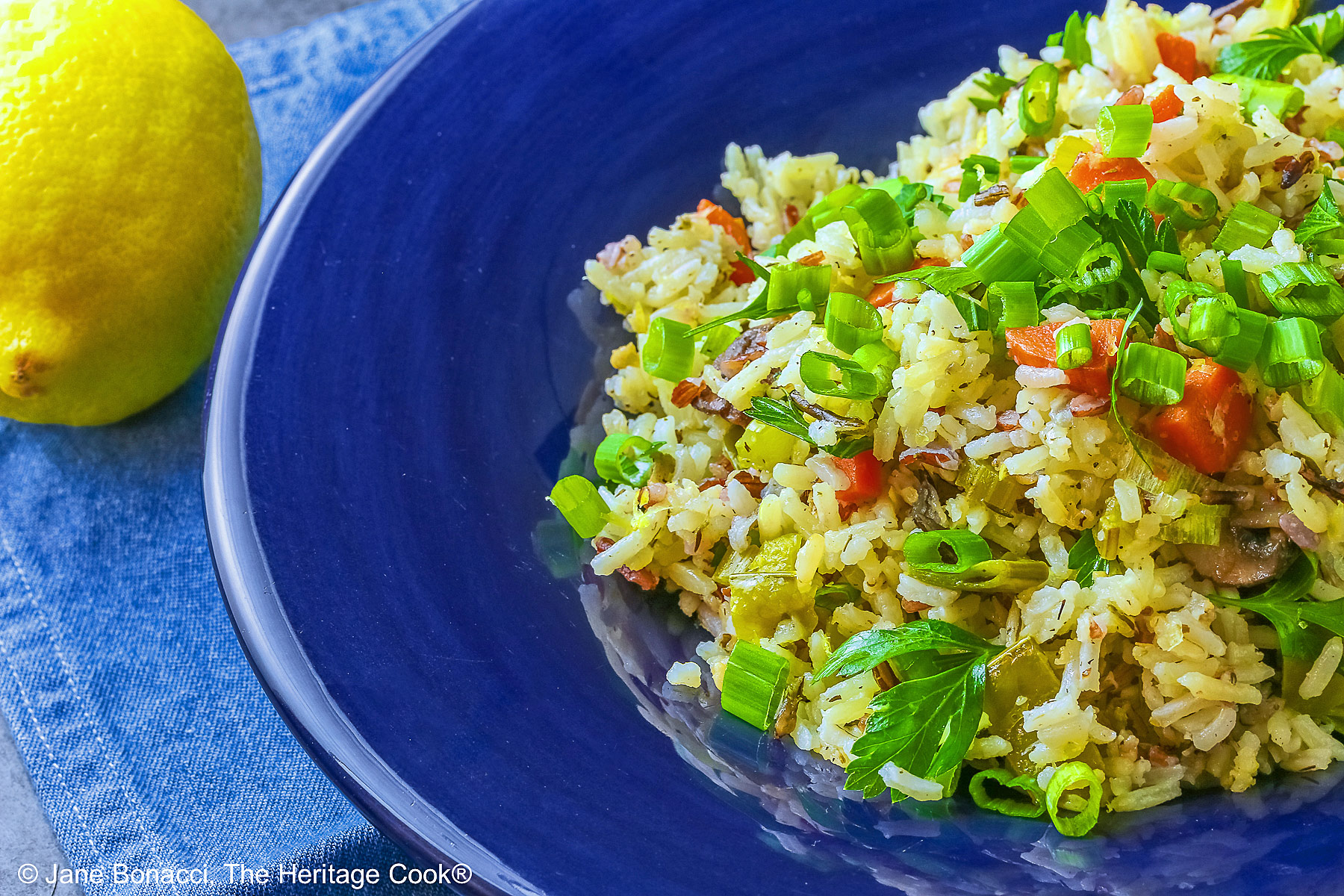 White and Wild Rice Pilaf with tons of vegetables is piled high in a deep blue bowl with a wide rim, topped with parsley and sliced green onions with three lemon slices along one edge © 2025 Jane Bonacci, The Heritage Cook.