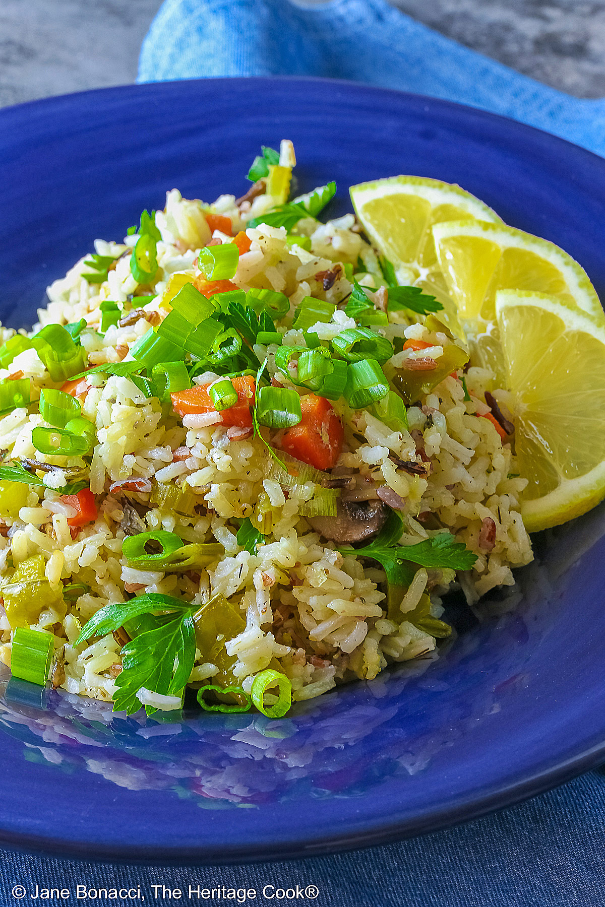 White and Wild Rice Pilaf with tons of vegetables is piled high in a deep blue bowl with a wide rim, topped with parsley and sliced green onions with three lemon slices along one edge © 2025 Jane Bonacci, The Heritage Cook.