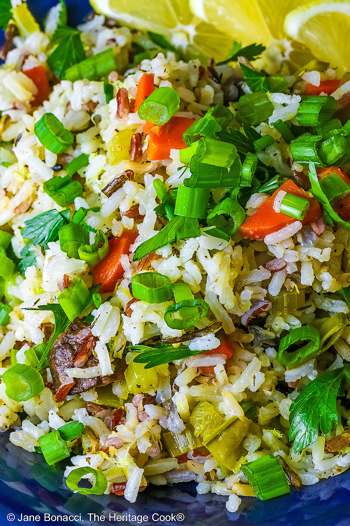 White and Wild Rice Pilaf with tons of vegetables is piled high in a deep blue bowl with a wide rim, topped with parsley and sliced green onions with three lemon slices along one edge © 2025 Jane Bonacci, The Heritage Cook.