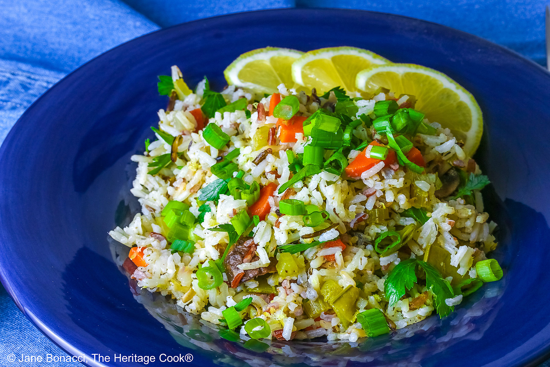 White and Wild Rice Pilaf with tons of vegetables is piled high in a deep blue bowl with a wide rim, topped with parsley and sliced green onions with three lemon slices along one edge © 2025 Jane Bonacci, The Heritage Cook.