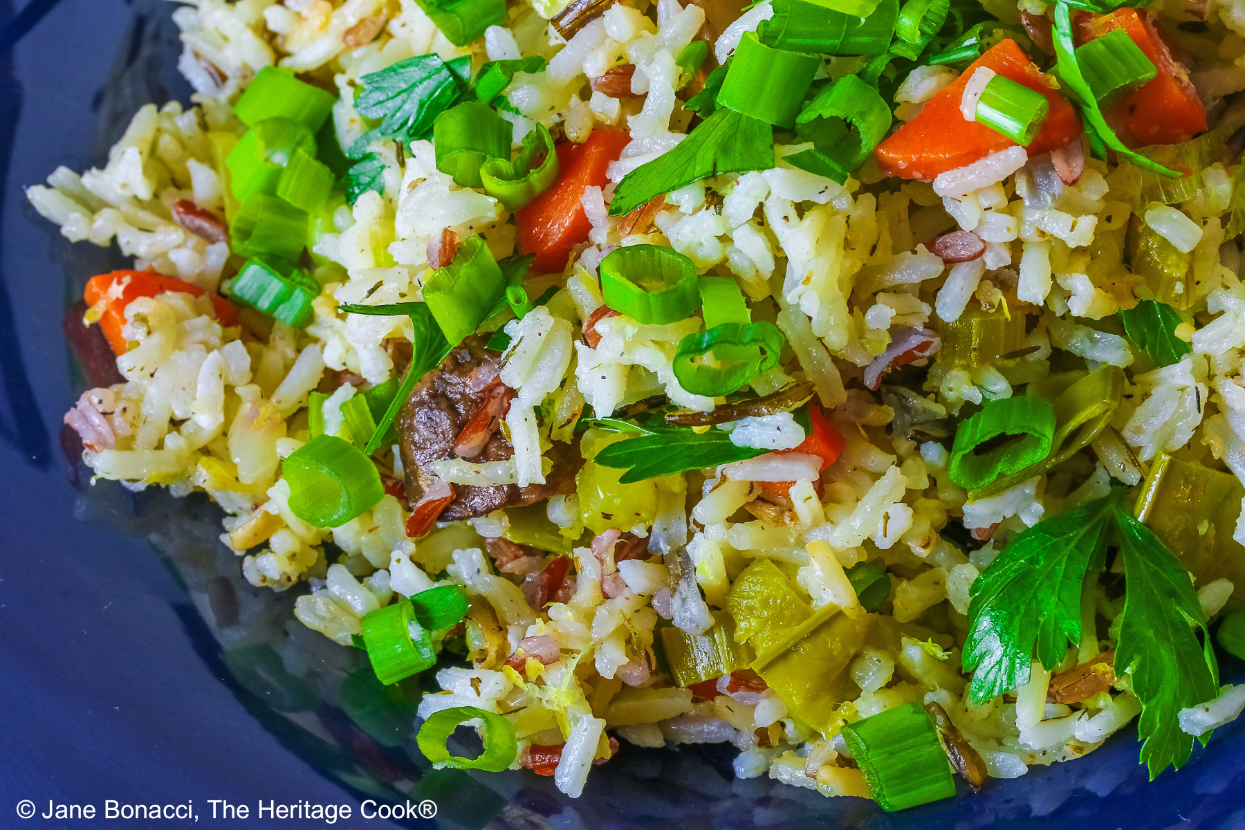 White and Wild Rice Pilaf with tons of vegetables is piled high in a deep blue bowl with a wide rim, topped with parsley and sliced green onions with three lemon slices along one edge © 2025 Jane Bonacci, The Heritage Cook.
