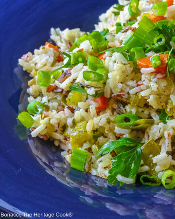 White and Wild Rice Pilaf with tons of vegetables is piled high in a deep blue bowl with a wide rim, topped with parsley and sliced green onions with three lemon slices along one edge © 2025 Jane Bonacci, The Heritage Cook.