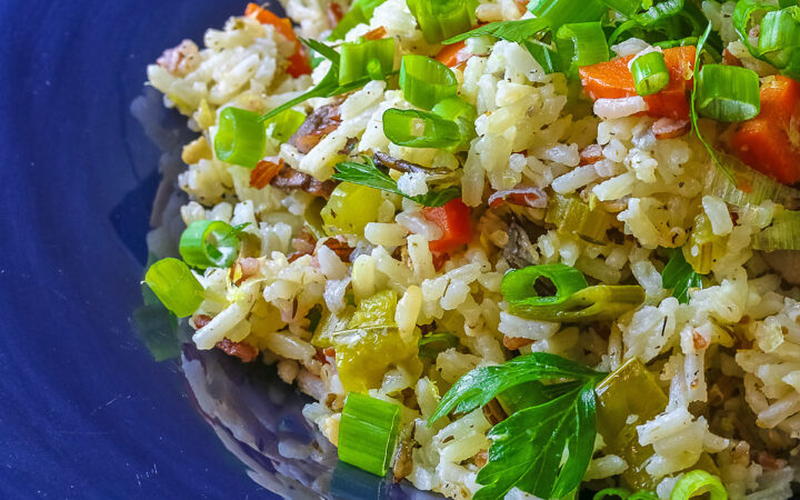 White and Wild Rice Pilaf with tons of vegetables is piled high in a deep blue bowl with a wide rim, topped with parsley and sliced green onions with three lemon slices along one edge © 2025 Jane Bonacci, The Heritage Cook.