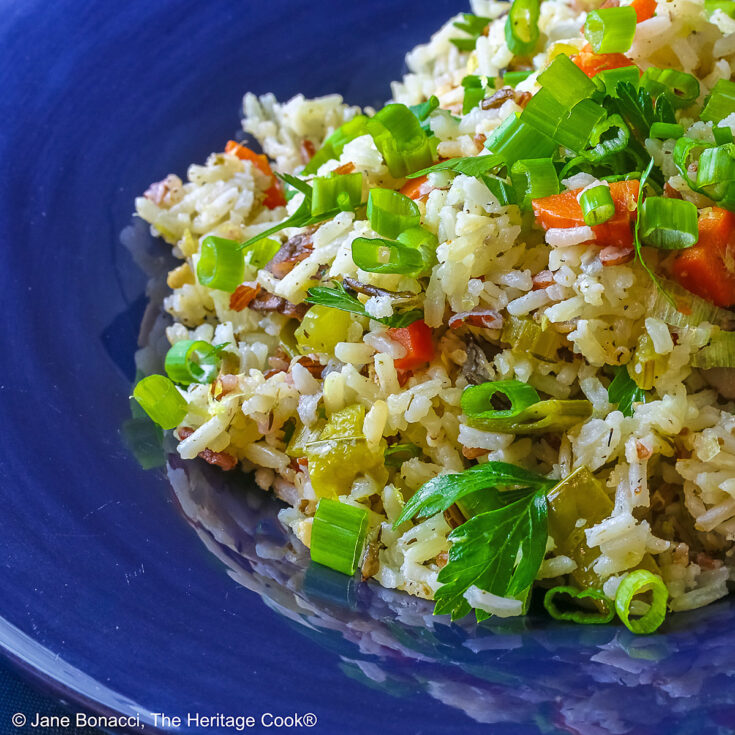White and Wild Rice Pilaf with tons of vegetables is piled high in a deep blue bowl with a wide rim, topped with parsley and sliced green onions with three lemon slices along one edge © 2025 Jane Bonacci, The Heritage Cook.