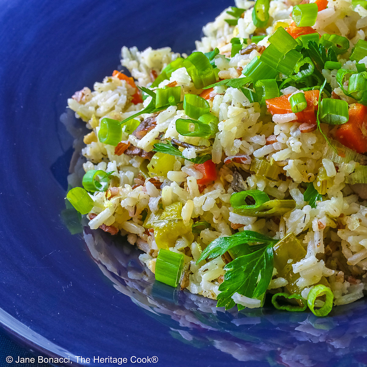 White and Wild Rice Pilaf with tons of vegetables is piled high in a deep blue bowl with a wide rim, topped with parsley and sliced green onions with three lemon slices along one edge © 2025 Jane Bonacci, The Heritage Cook.