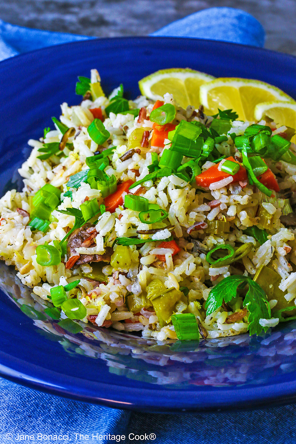 White and Wild Rice Pilaf with tons of vegetables is piled high in a deep blue bowl with a wide rim, topped with parsley and sliced green onions with three lemon slices along one edge © 2025 Jane Bonacci, The Heritage Cook.