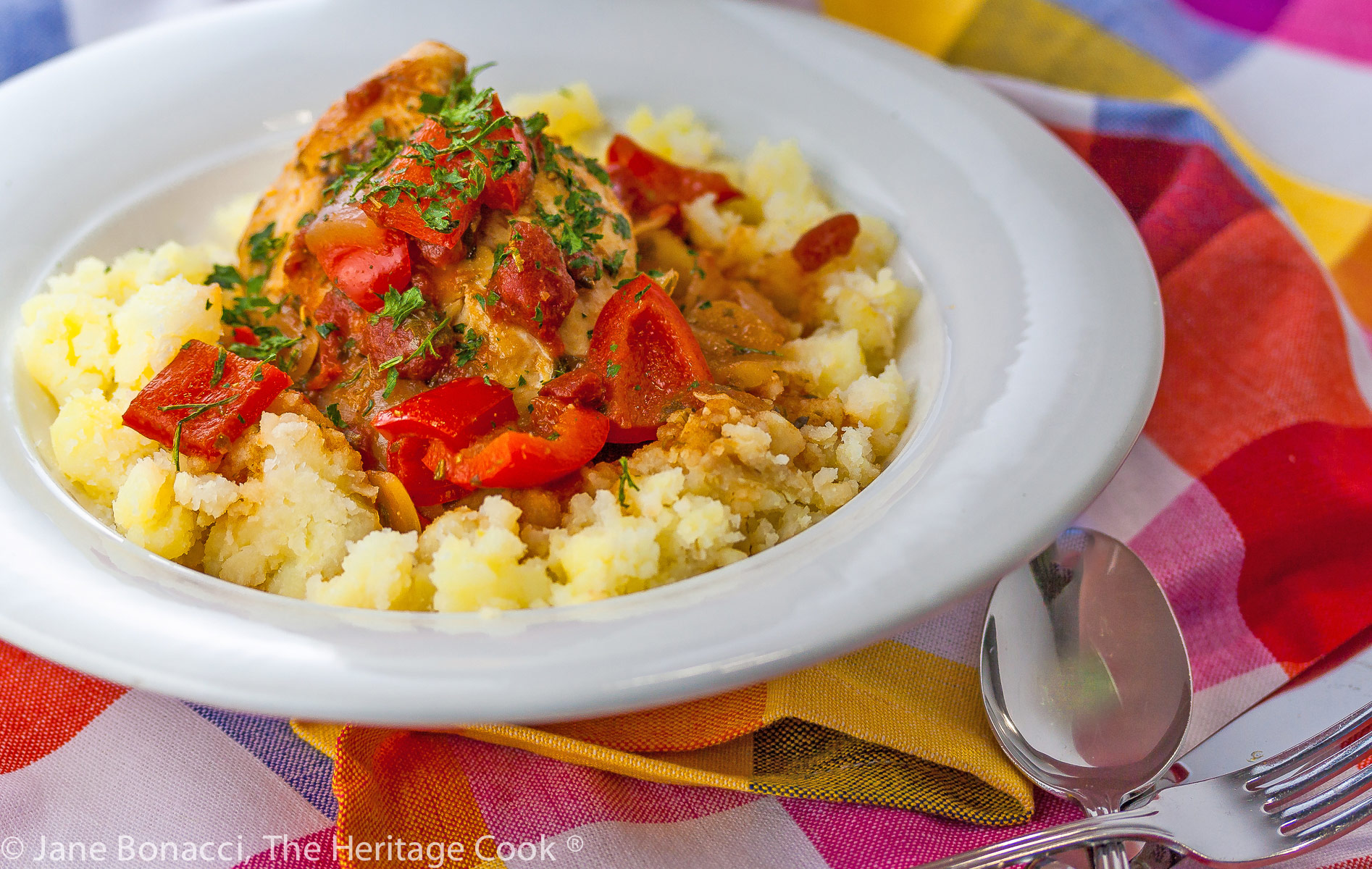 A white bowl with smashed yellow Yukon Gold potatoes is topped with chicken braised in tomatoes, onions, mushrooms, and wine; Comforting Chicken Cacciatore with smashed Yukon Gold potatoes © 2023 Jane Bonacci, The Heritage Cook. 