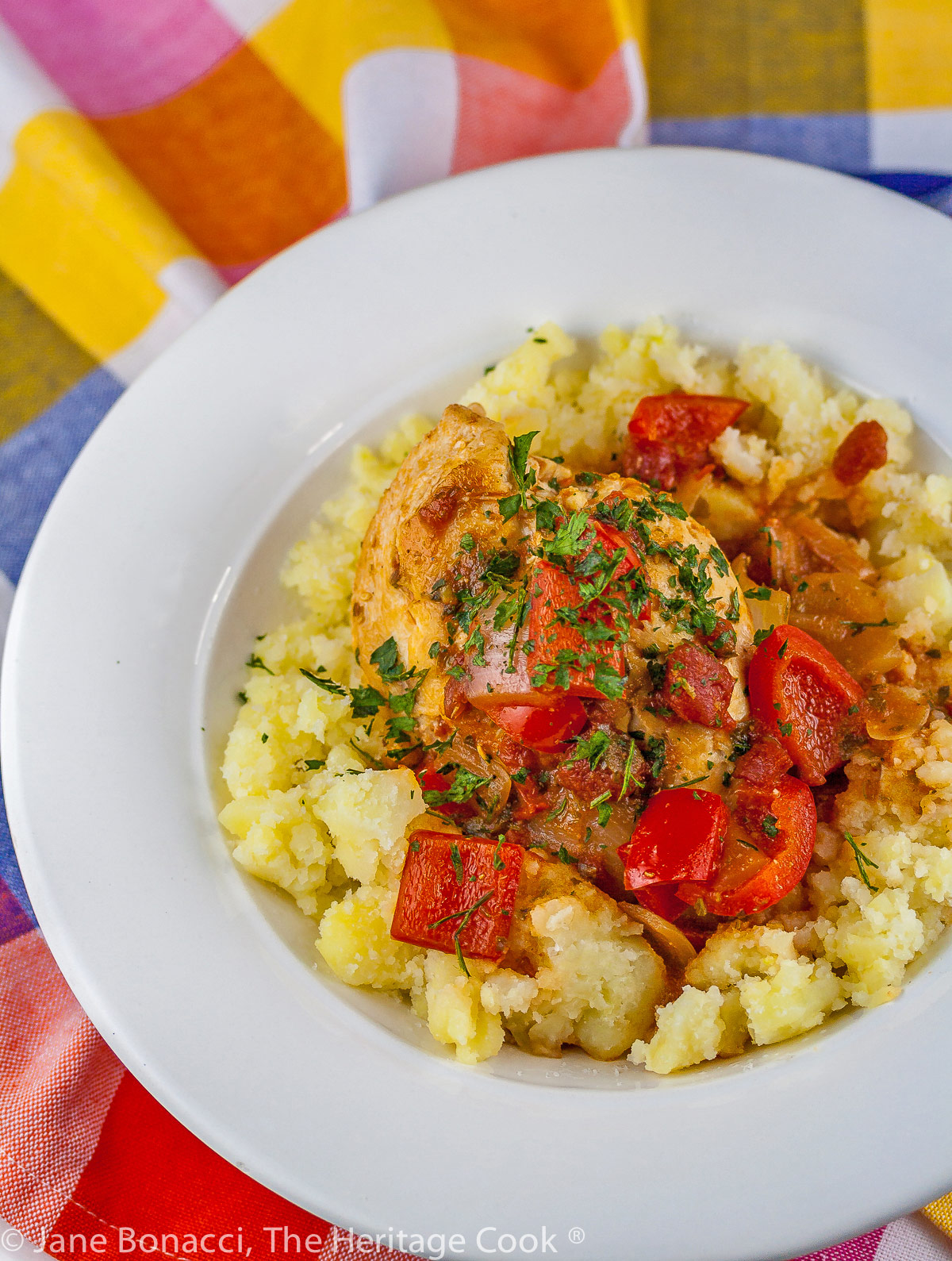 A white bowl with smashed yellow Yukon Gold potatoes is topped with chicken braised in tomatoes, onions, mushrooms, and wine; Comforting Chicken Cacciatore with smashed Yukon Gold potatoes © 2023 Jane Bonacci, The Heritage Cook. 