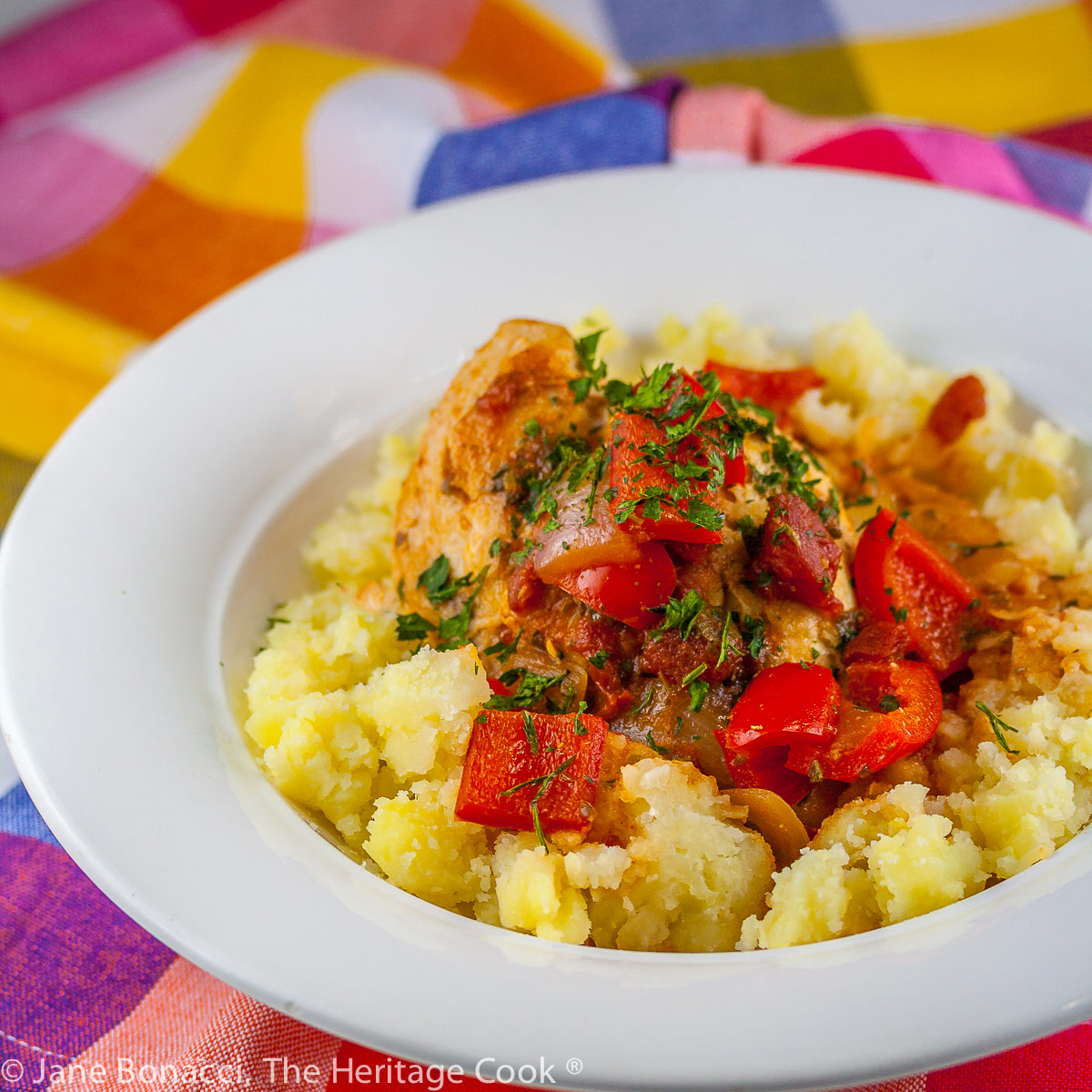 A white bowl with smashed yellow Yukon Gold potatoes is topped with chicken braised in tomatoes, onions, mushrooms, and wine; Comforting Chicken Cacciatore with smashed Yukon Gold potatoes © 2023 Jane Bonacci, The Heritage Cook.