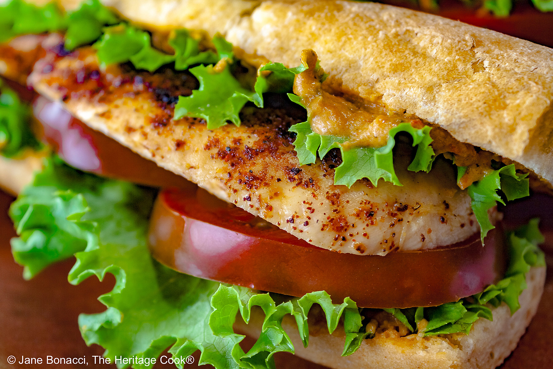 One or two chicken sandwiches with hummus aioli, rolls stuffed with grilled chicken breasts, lettuce, tomatoes, hummus aioli, sitting on a colorful plaid cloth with a deep blue background behind © 2025 Jane Bonacci, The Heritage Cook. 