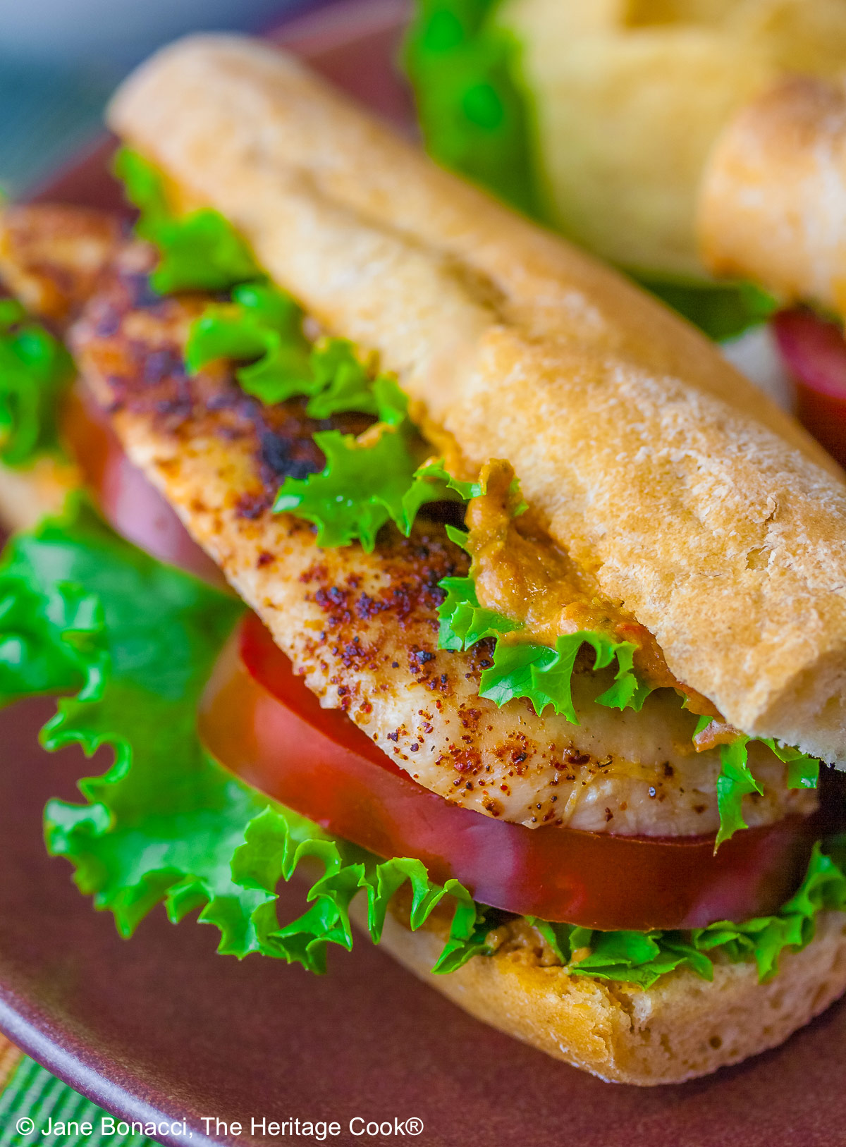 One or two chicken sandwiches with hummus aioli, rolls stuffed with grilled chicken breasts, lettuce, tomatoes, hummus aioli, sitting on a colorful plaid cloth with a deep blue background behind © 2025 Jane Bonacci, The Heritage Cook. 