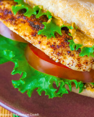 One or two chicken sandwiches with hummus aioli, rolls stuffed with grilled chicken breasts, lettuce, tomatoes, hummus aioli, sitting on a colorful plaid cloth with a deep blue background behind © 2025 Jane Bonacci, The Heritage Cook.