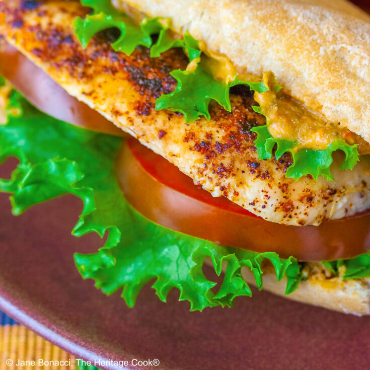 One or two chicken sandwiches with hummus aioli, rolls stuffed with grilled chicken breasts, lettuce, tomatoes, hummus aioli, sitting on a colorful plaid cloth with a deep blue background behind © 2025 Jane Bonacci, The Heritage Cook.