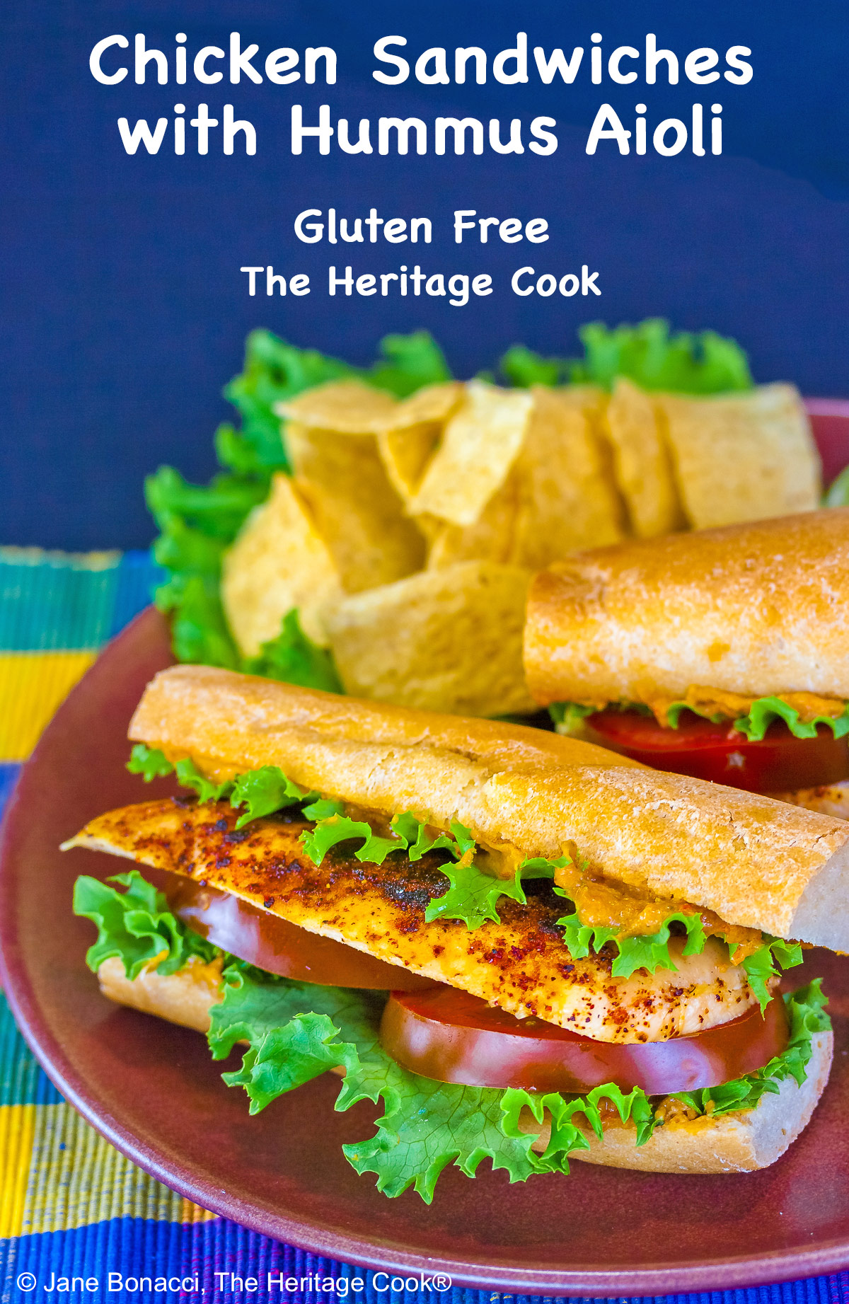 One or two chicken sandwiches with hummus aioli, rolls stuffed with grilled chicken breasts, lettuce, tomatoes, hummus aioli, sitting on a colorful plaid cloth with a deep blue background behind © 2025 Jane Bonacci, The Heritage Cook. 