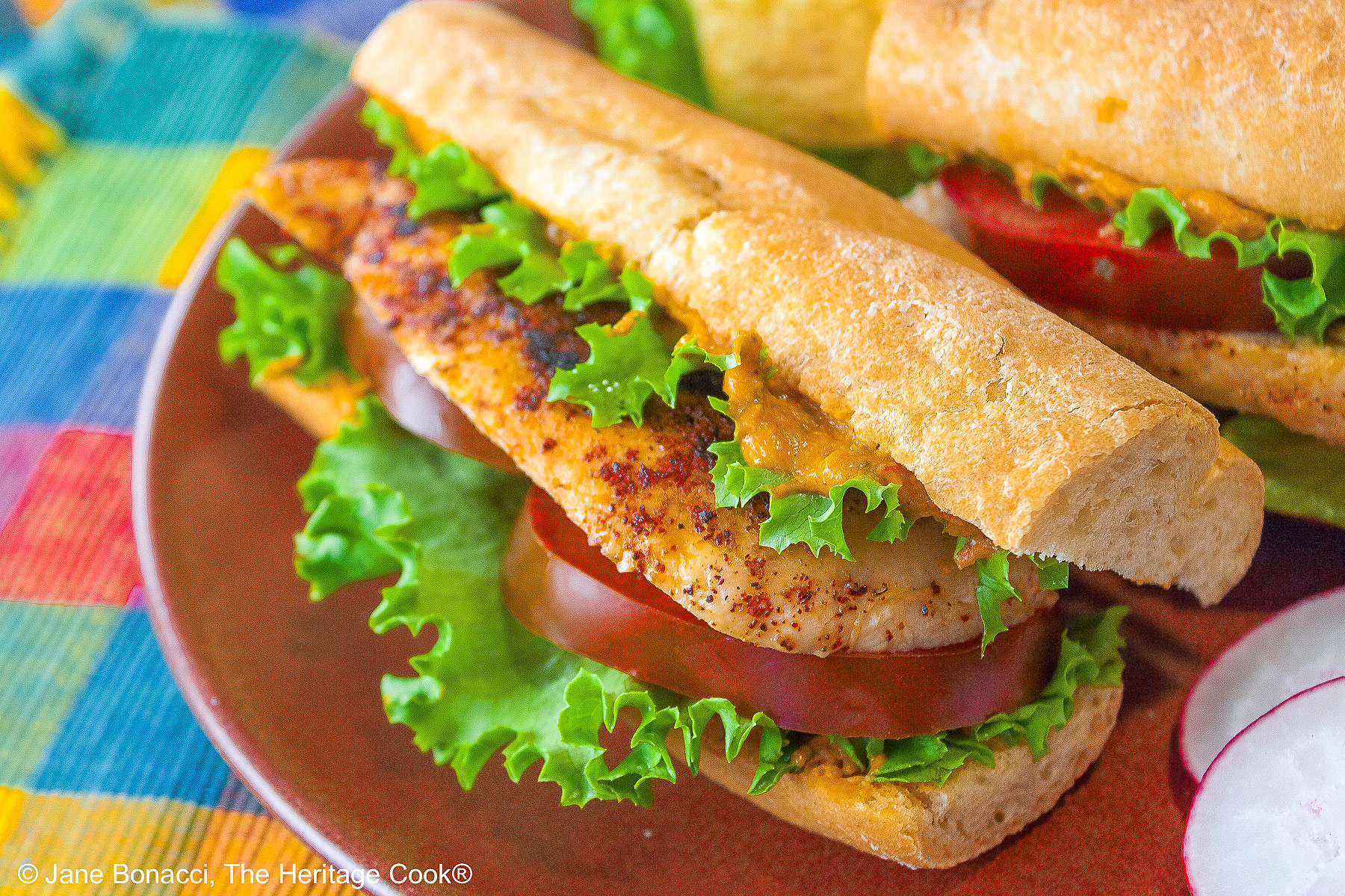 One or two chicken sandwiches with hummus aioli, rolls stuffed with grilled chicken breasts, lettuce, tomatoes, hummus aioli, sitting on a colorful plaid cloth with a deep blue background behind © 2025 Jane Bonacci, The Heritage Cook. 