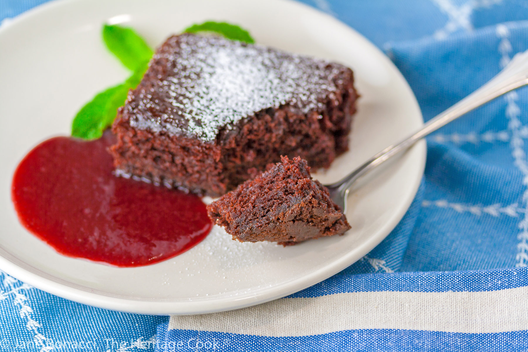 Square slice of chocolate cake surrounded by red raspberry sauce on a white plate with a mint sprig on blue background; Luscious Chocolate Cake with Raspberry Sauce © 2023 Jane Bonacci, The Heritage Cook.