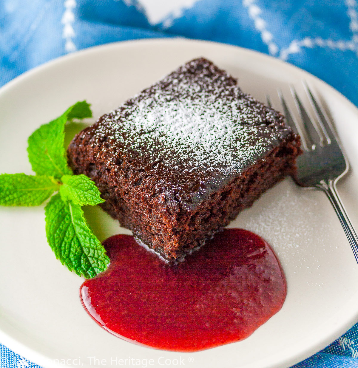 Square slice of chocolate cake surrounded by red raspberry sauce on a white plate with a mint sprig on blue background; Luscious Chocolate Cake with Raspberry Sauce © 2023 Jane Bonacci, The Heritage Cook.