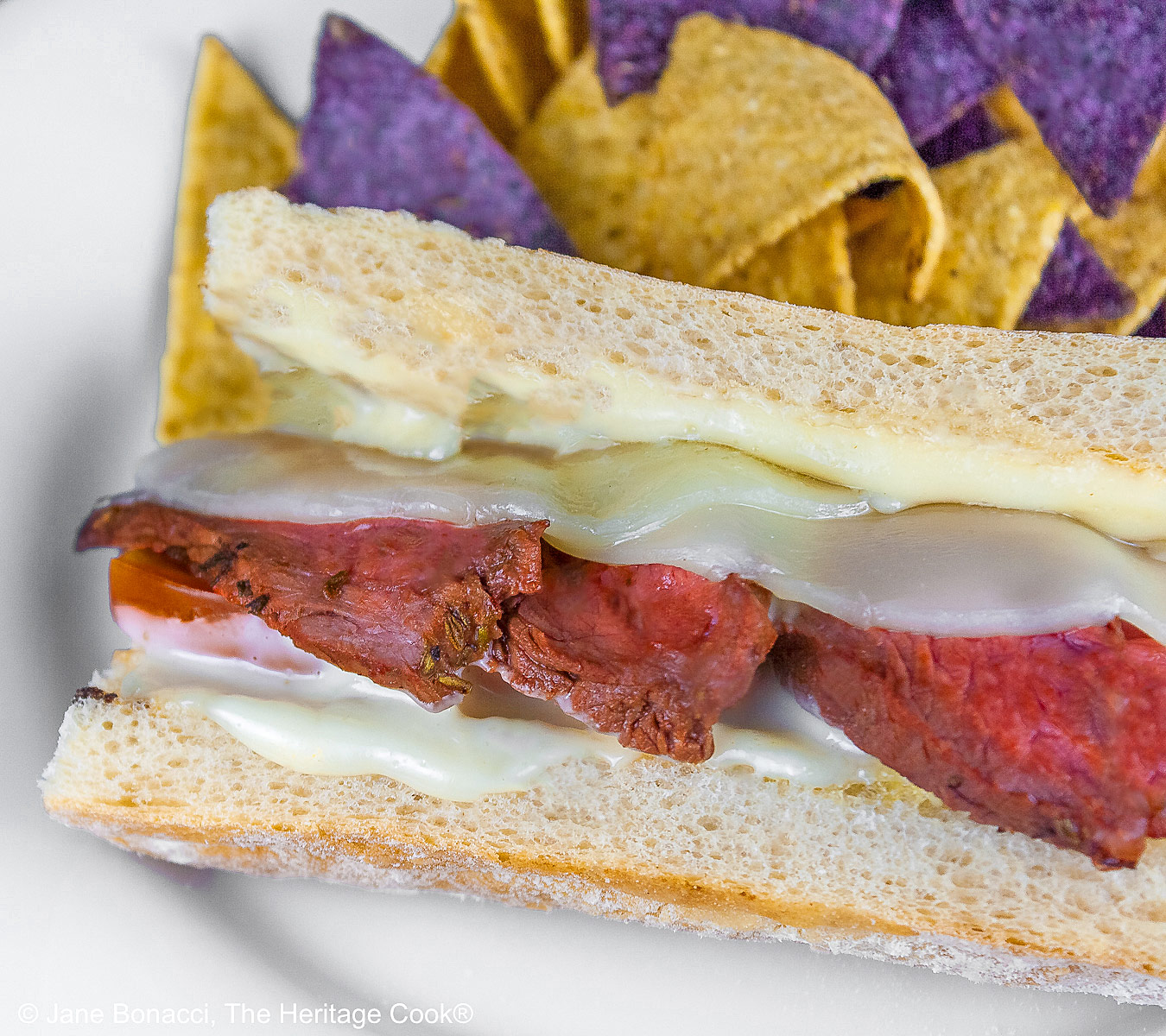 Large split sandwich roll stuffed with grilled sliced steak and melted Provolone cheese on a white plate, flanked by yellow and blue corn chips and some with sliced tomatoes, sitting on burlap and brightly colored checked cloth © 2025 Jane Bonacci, The Heritage Cook. 