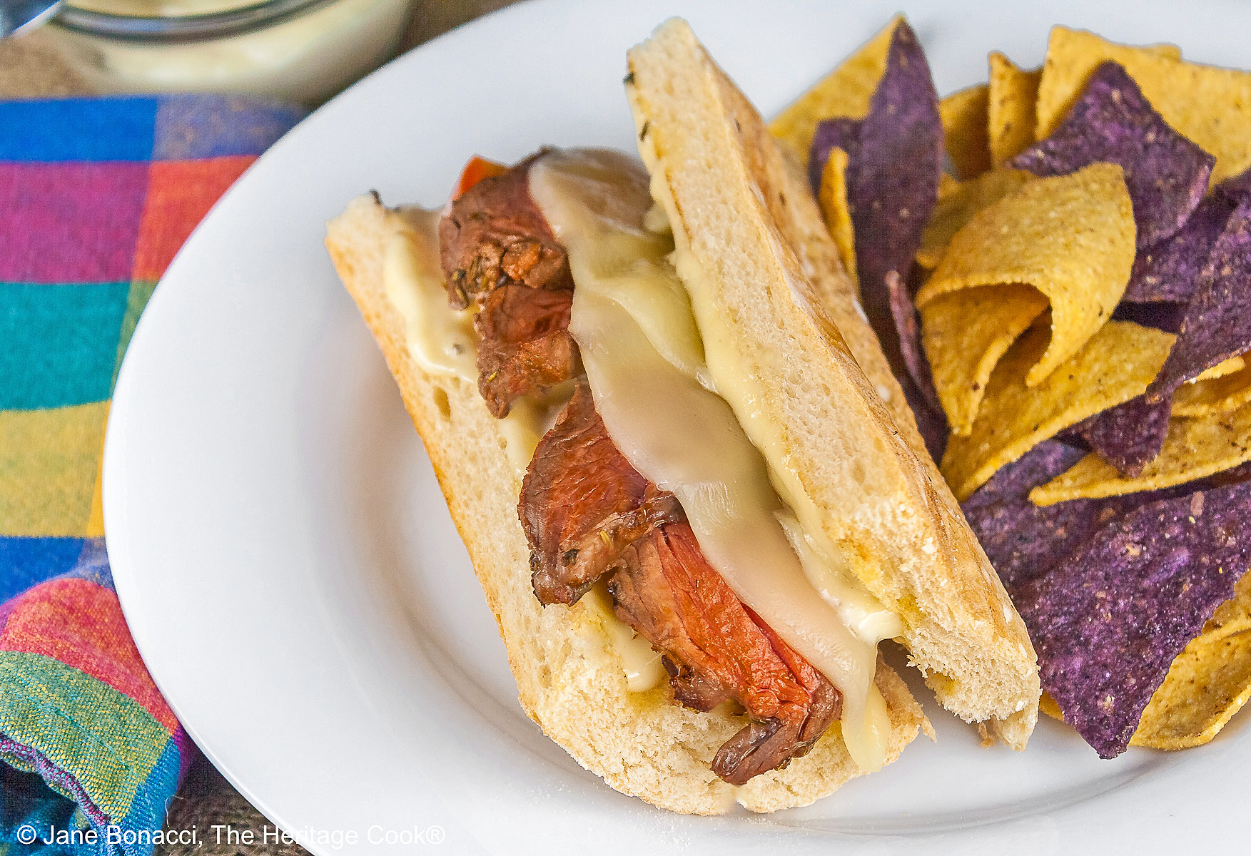 Large split sandwich roll stuffed with grilled sliced steak and melted Provolone cheese on a white plate, flanked by yellow and blue corn chips and some with sliced tomatoes, sitting on burlap and brightly colored checked cloth © 2025 Jane Bonacci, The Heritage Cook. 