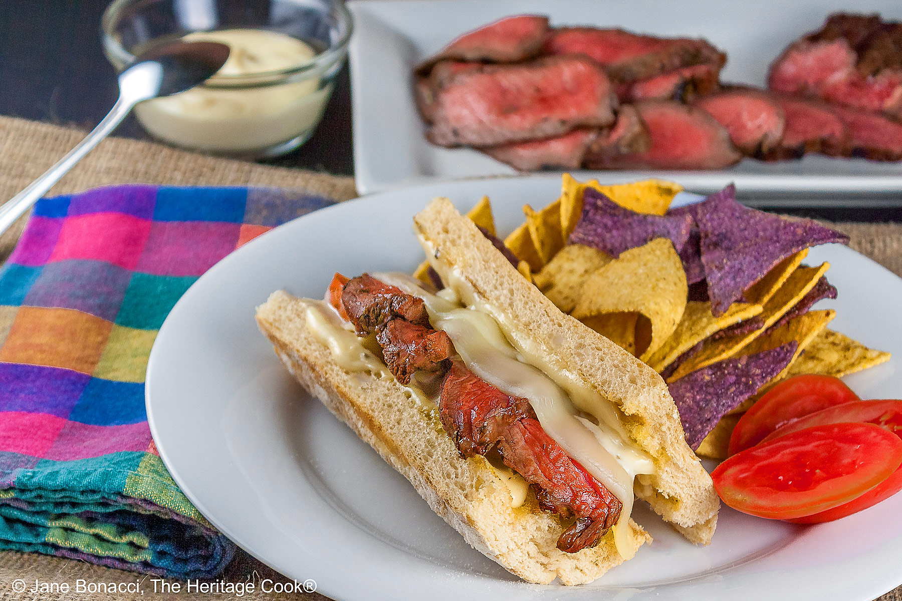 Large split sandwich roll stuffed with grilled sliced steak and melted Provolone cheese on a white plate, flanked by yellow and blue corn chips and some with sliced tomatoes, sitting on burlap and brightly colored checked cloth © 2025 Jane Bonacci, The Heritage Cook. 