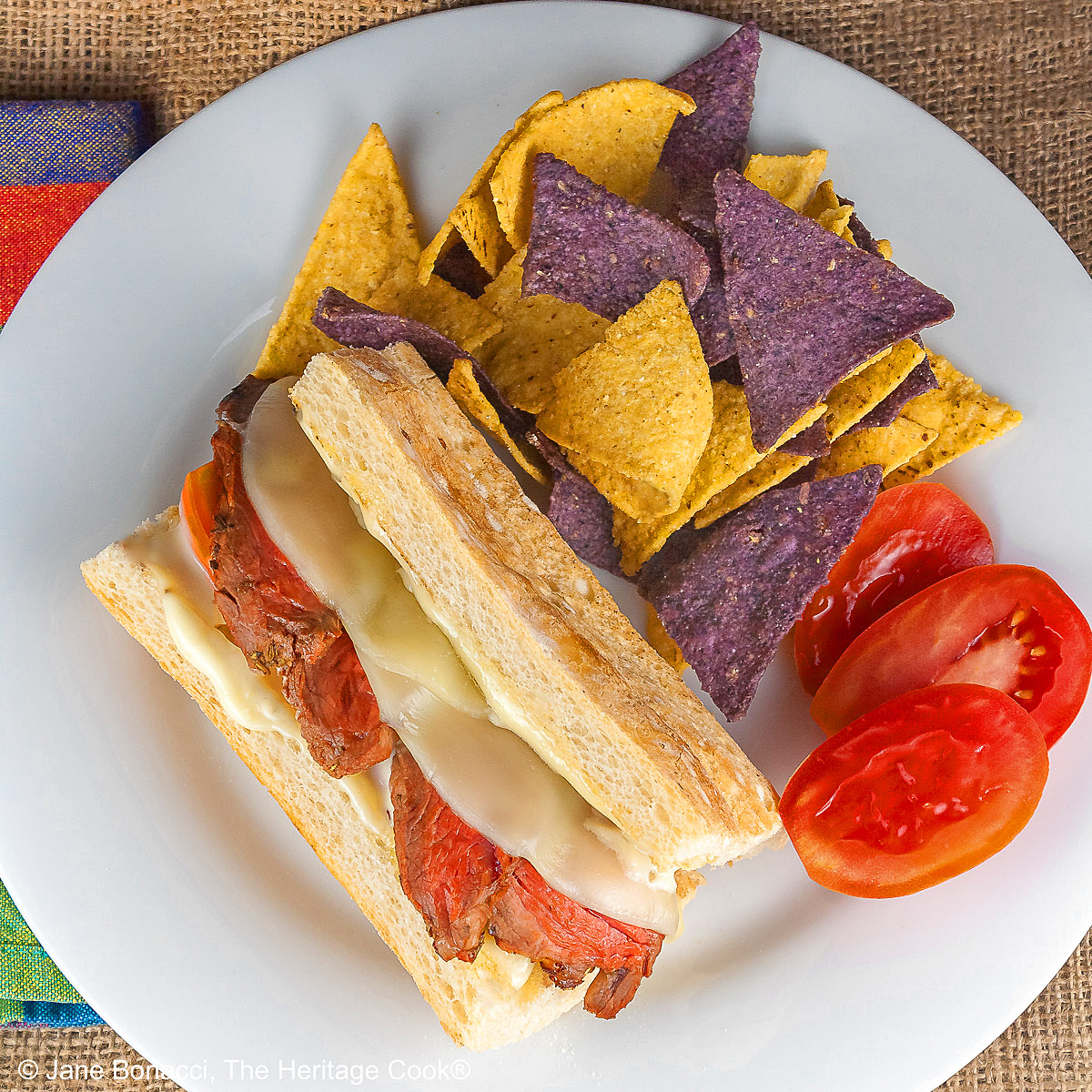 Large split sandwich roll stuffed with grilled sliced steak and melted Provolone cheese on a white plate, flanked by yellow and blue corn chips and some with sliced tomatoes, sitting on burlap and brightly colored checked cloth © 2025 Jane Bonacci, The Heritage Cook. 