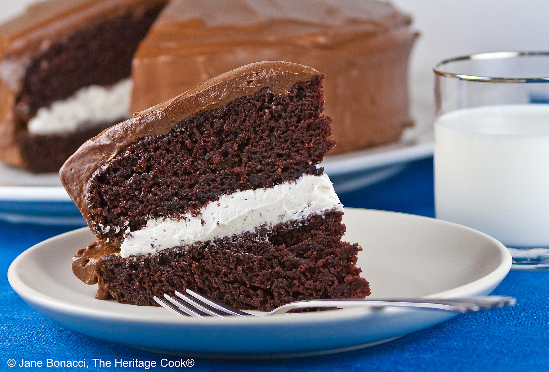 Slice of the Little Debbie Chocolate Layer Cake with vanilla filling on a white plate with a fork beside it, with a glass of milk behind and the whole cake with the slice cut out in the background © 2026 Jane Bonacci, The Heritage Cook.