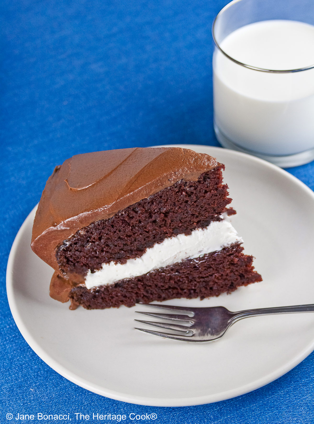 Slice of the Little Debbie Chocolate Layer Cake with vanilla filling on a white plate with a fork beside it, with a glass of milk behind and the whole cake with the slice cut out in the background © 2026 Jane Bonacci, The Heritage Cook.