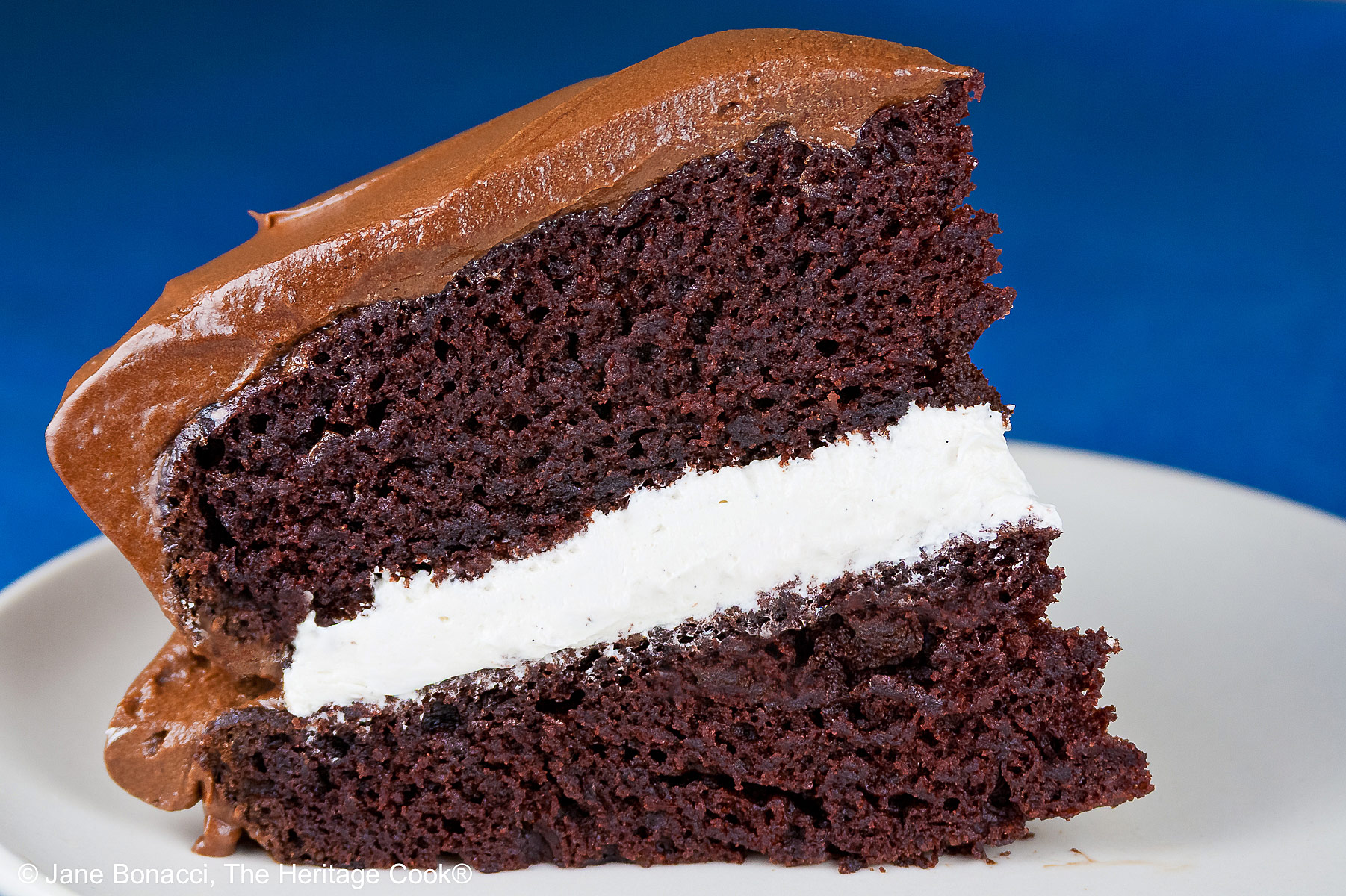Slice of the Little Debbie Chocolate Layer Cake with vanilla filling on a white plate with a fork beside it, with a glass of milk behind and the whole cake with the slice cut out in the background © 2026 Jane Bonacci, The Heritage Cook.