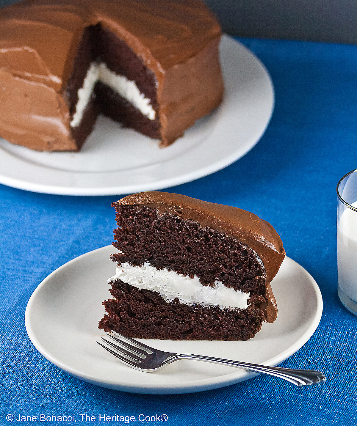 Slice of the Little Debbie Chocolate Layer Cake with vanilla filling on a white plate with a fork beside it, with a glass of milk behind and the whole cake with the slice cut out in the background © 2026 Jane Bonacci, The Heritage Cook.